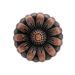 May include: A round metal cabinet knob featuring a raised floral design with a copper-toned finish. The center of the flower has a textured, dotted surface, surrounded by radiating petals with dark, recessed lines between each petal segment.