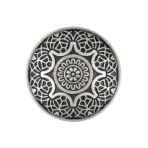 Pode incluir: Um botão circular com um acabamento metálico prateado em relevo sobre um fundo preto rebaixado. O design apresenta um motivo floral central rodeado por um anel de pétalas, envolvido por um intrincado padrão de grelha geométrica.