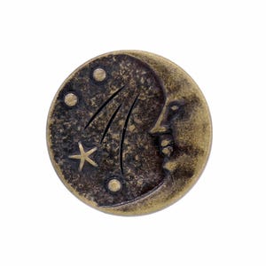 May include: A round, antique brass-toned button featuring a relief design of a crescent moon with a human-like face. The surface includes three small circular indentations and a five-pointed star, all set against a dark, textured background.