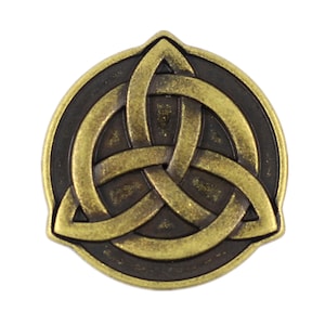 Può includere: Una spilla metallica circolare con finitura in ottone anticato e un design a nodo triquetra in rilievo. Il motivo centrale intrecciato è posto su uno sfondo marrone scuro incassato, creando un netto contrasto tra le linee metalliche color oro e le aree in ombra dell'emblema.