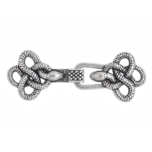 Op de afbeelding: Een zilverkleurige metalen sluiting voor een mantel met twee symmetrische slangenmotieven. Elke slang is gevormd in een ingewikkeld knoppatroon met gedetailleerde schubben. De twee helften zijn verbonden door een centrale rechthoekige metalen lus.