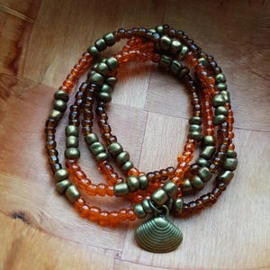Peut inclure: Plusieurs bracelets de perles aux tons orange, marron et bronze. Les bracelets sont composés de petites perles rondes et de perles carrées plus grandes. Un charm en forme de coquillage en bronze est suspendu.