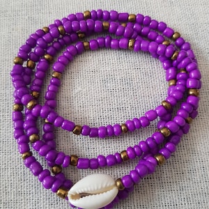 Peut inclure: Une chaîne de taille à perles violettes superposées avec des accents dorés et un pendentif en coquillage blanc. Les perles sont petites et rondes, créant un aspect texturé. La chaîne de taille est présentée sur un fond neutre.