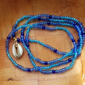Peut inclure: Un ensemble de perles de taille bleues avec un pendentif coquillage blanc et doré. Les perles sont de différentes nuances de bleu, créant un effet superposé. Le pendentif coquillage ajoute une touche unique.