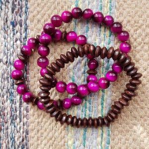 Peut inclure: Trois bracelets : deux avec des perles magenta vives et un avec des perles en bois brun foncé. Les bracelets sont disposés sur un fond de tissu texturé de tons neutres. Les perles sont rondes et lisses, créant un contraste de texture.
