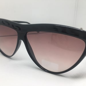 Puede incluir: Gafas de sol negras con forma de ojo de gato y lentes degradadas. Las monturas tienen un diseño decorativo en la parte superior. Las lentes hacen una transición de rosa claro a un tono más oscuro.