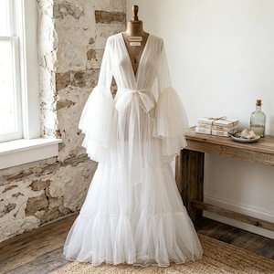 May include: A floor-length white sheer robe featuring a deep V-neckline, long tiered bell sleeves, and a matching fabric tie at the waist. The garment is made of a lightweight, translucent tulle fabric and is displayed on a dress form mannequin.