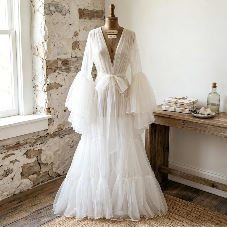 May include: A floor-length white sheer robe featuring a deep V-neckline, long tiered bell sleeves, and a matching fabric tie at the waist. The garment is made of a lightweight, translucent tulle fabric and is displayed on a dress form mannequin.