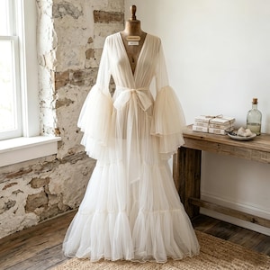 May include: A floor-length, off-white sheer tulle robe featuring a deep V-neckline, long tiered bell sleeves, and a matching fabric tie belt at the waist. The garment is displayed on a dress form, showcasing a layered, flowing skirt design.