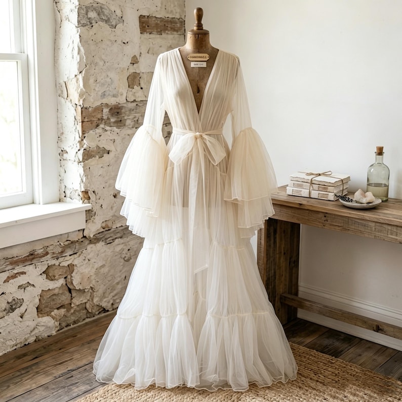 May include: A floor-length, off-white sheer tulle robe featuring a deep V-neckline, long tiered bell sleeves, and a matching fabric tie belt at the waist. The garment is displayed on a dress form, showcasing a layered, flowing skirt design.