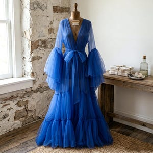 May include: A floor-length royal blue robe made of sheer tulle fabric. The garment features a deep V-neckline, long voluminous bell sleeves with tiered ruffles, and a matching tie belt at the waist. The skirt is constructed with multiple layers.