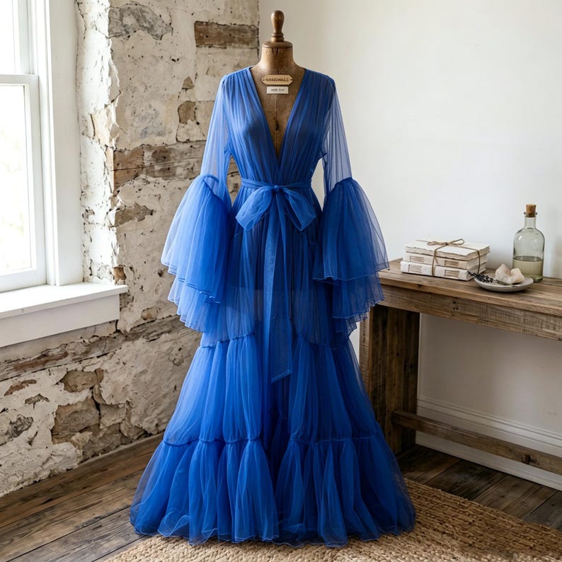 May include: A floor-length royal blue robe made of sheer tulle fabric. The garment features a deep V-neckline, long voluminous bell sleeves with tiered ruffles, and a matching tie belt at the waist. The skirt is constructed with multiple layers.