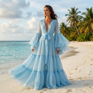 May include: A floor-length light blue dress featuring a deep V-neckline, long sheer bell sleeves with ruffled cuffs, and a tiered skirt. The dress includes a matching fabric tie at the waist, creating a gathered effect in the bodice and skirt.