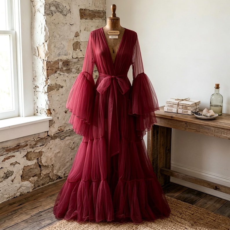 May include: A floor-length burgundy robe made of sheer, layered tulle fabric. The garment features a deep V-neckline, long voluminous bell sleeves with tiered ruffles, and a matching fabric tie belt at the waist to secure the front closure.