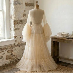 May include: A floor-length, cream-colored tulle gown displayed on a mannequin. The dress features long, voluminous bell sleeves, a fitted bodice with a defined waistline, and a multi-tiered, gathered skirt that flows down to the floor.