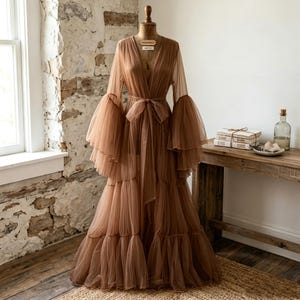 May include: A floor-length, sheer brown tulle robe featuring long, voluminous bell sleeves with tiered ruffles, a deep V-neckline, and a matching fabric tie belt at the waist. The garment is displayed on a dress form, showcasing its layered skirt.