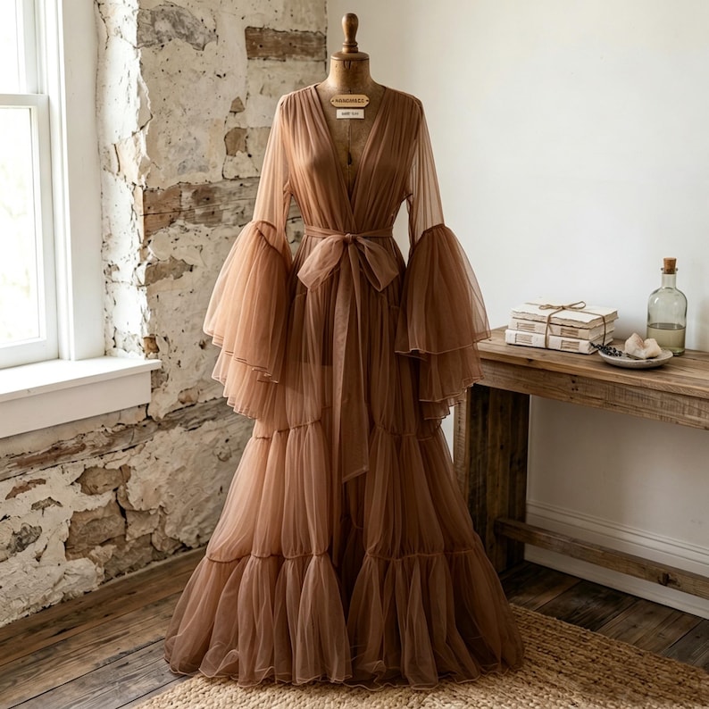 May include: A floor-length, sheer brown tulle robe featuring long, voluminous bell sleeves with tiered ruffles, a deep V-neckline, and a matching fabric tie belt at the waist. The garment is displayed on a dress form, showcasing its layered skirt.