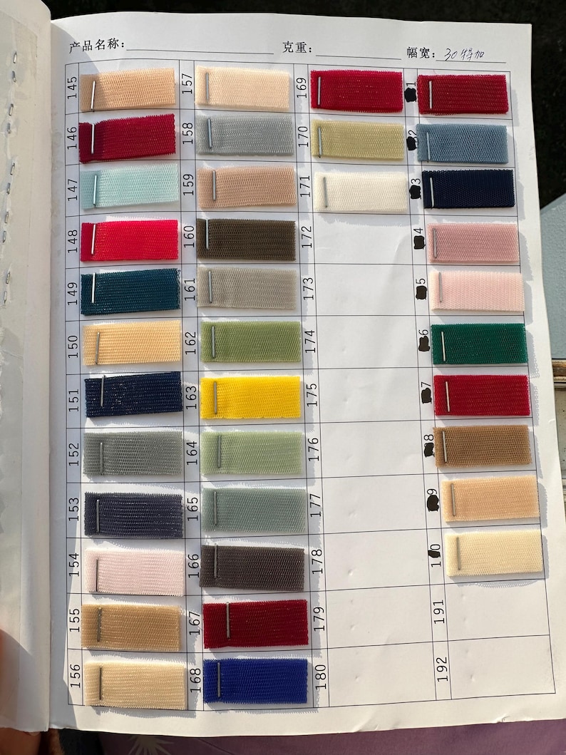 May include: A fabric swatch color chart featuring a grid of numbered rows and columns. Each cell contains a small rectangular piece of mesh fabric in various colors, including red, blue, green, yellow, beige, and gray, attached with a metal staple.