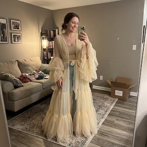 May include: A floor-length, sheer beige tulle duster robe featuring voluminous, multi-tiered ruffled sleeves and a matching tie-waist closure. The garment is worn over a simple beige tank top and light blue denim jeans, creating a layered look.