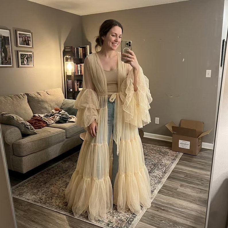 May include: A floor-length, sheer beige tulle duster robe featuring voluminous, multi-tiered ruffled sleeves and a matching tie-waist closure. The garment is worn over a simple beige tank top and light blue denim jeans, creating a layered look.