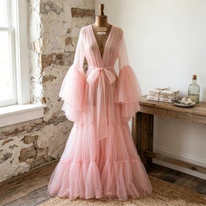 May include: A floor-length, pale pink sheer tulle robe featuring long, voluminous tiered bell sleeves and a deep V-neckline. The garment includes a matching fabric tie belt at the waist and a multi-layered, ruffled skirt that flows to the floor.