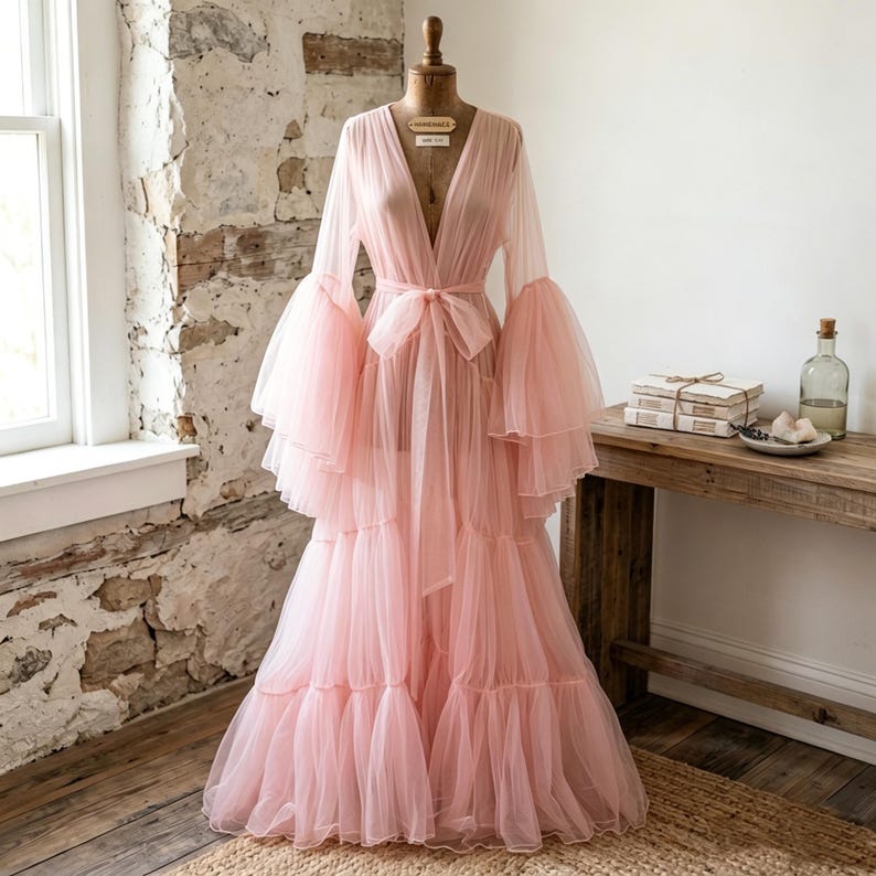 May include: A floor-length, pale pink sheer tulle robe featuring long, voluminous tiered bell sleeves and a deep V-neckline. The garment includes a matching fabric tie belt at the waist and a multi-layered, ruffled skirt that flows to the floor.