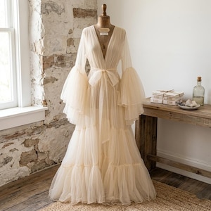 May include: A cream-colored, long tulle robe with a plunging neckline and tiered skirt is displayed on a wooden dress form. The robe features long, flared sleeves and a matching tie belt. The background includes a rustic brick wall and a wooden table.