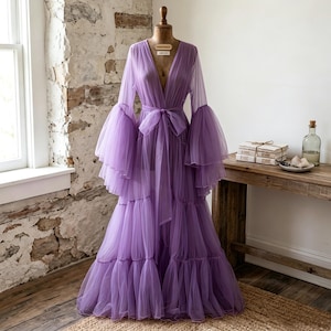May include: A floor-length lavender tulle robe featuring a deep V-neckline, long voluminous bell sleeves with tiered ruffles, and a matching fabric tie belt at the waist. The garment is displayed on a dress form and has a layered skirt design.