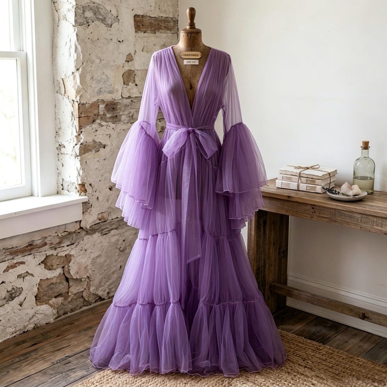 May include: A floor-length lavender tulle robe featuring a deep V-neckline, long voluminous bell sleeves with tiered ruffles, and a matching fabric tie belt at the waist. The garment is displayed on a dress form and has a layered skirt design.