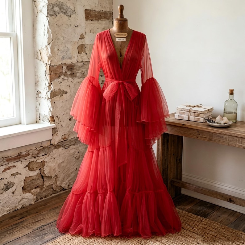 May include: A floor-length red robe made of sheer tulle fabric, featuring a deep V-neckline, long voluminous bell sleeves, and a matching tie belt at the waist. The garment has a tiered skirt design with gathered layers that create a full silhouette.