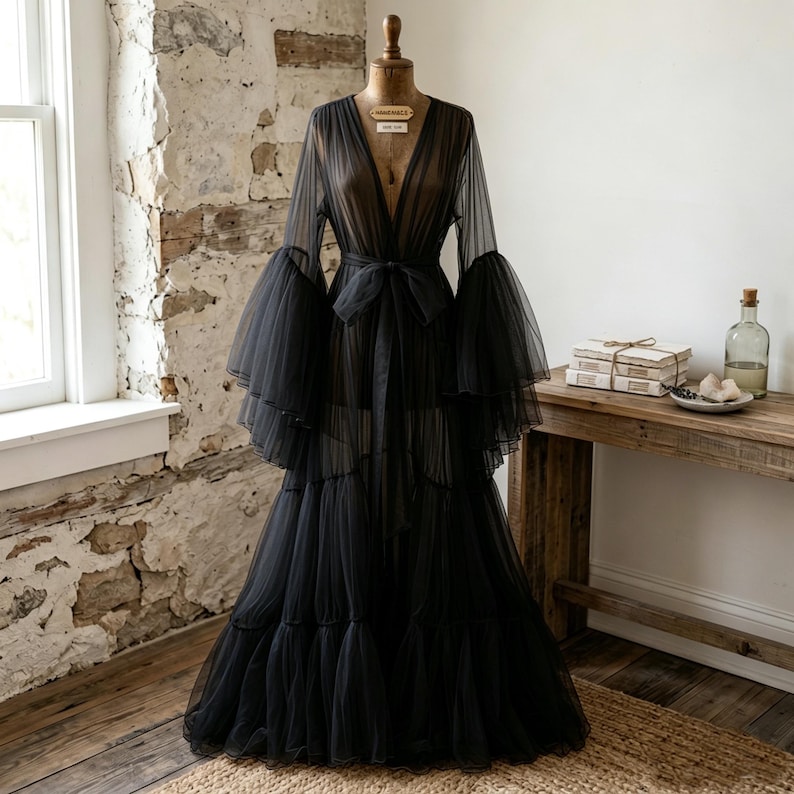 May include: A floor-length black sheer tulle robe featuring a deep V-neckline, long voluminous bell sleeves with tiered ruffles, and a matching fabric tie belt at the waist. The garment is displayed on a dress form, showcasing its layered design.