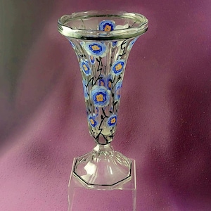 Könnte beinhalten: Eine klare Glasvase mit ausgestelltem Oberteil und sechseckigem Sockel. Die Vase ist mit blau-gelben Blumendesigns und schwarzen Stielen verziert. Der Rand der Vase hat eine silberne Zierleiste. Die Vase wird auf einem transparenten Ständer ausgestellt.