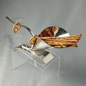 Op de afbeelding: Een grillig zilverkleurig serveerschaaltje voor pretzels in de vorm van een bezem. De bezem bevat een bundel pretzelstokjes en een pretzelring aan het uiteinde van de steel. De schaal wordt gepresenteerd op een heldere acrylstandaard.