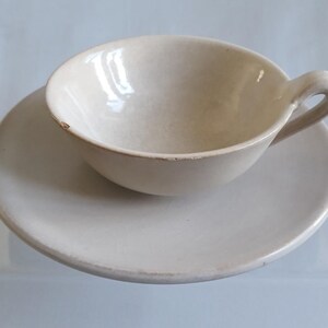 Könnte beinhalten: Ein cremefarbenes Keramik-Tassen- und Untertassen-Set. Die Tasse hat eine abgerundete Form mit einem Henkel, und die Untertasse ist kreisförmig. Das Set hat ein rustikales, handgemachtes Aussehen mit leichten Unvollkommenheiten.