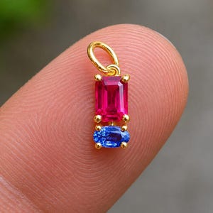 May include: A gold pendant featuring a rectangular, vibrant pink gemstone above an oval, deep blue gemstone. The pendant is small, with a loop at the top for a chain. The gemstones are set in a gold setting.
