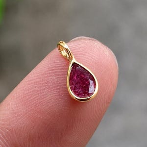 May include: A small teardrop-shaped pendant featuring a faceted, deep pink gemstone set in a polished yellow gold bezel. The pendant includes a small, integrated gold jump ring at the top for attachment to a chain or cord, showcasing a minimalist design.