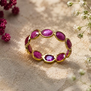 May include: A gold band ring featuring a continuous row of faceted, oval-shaped magenta-pink gemstones set in individual gold bezels. The ring is positioned on a textured surface, surrounded by small clusters of white and deep pink flowers.