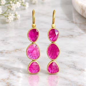 Puede incluir: Un par de pendientes colgantes con tres piedras preciosas de color rosa vibrante, facetadas y alineadas verticalmente en metal dorado. Cada pendiente cuelga de un sencillo gancho dorado, mostrando un acabado pulido y formas orgánicas.