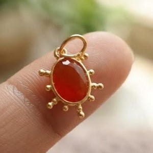 Pode incluir: Um pingente de tom dourado com uma pedra preciosa central de formato oval em um tom vermelho-alaranjado profundo. A estrutura de metal é adornada com pequenas esferas espaçadas uniformemente ao redor da borda e um aro circular no topo.