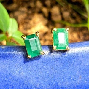 Puede incluir: Un par de pendientes de botón de color verde esmeralda con piedras de corte rectangular engastadas en garras de metal dorado. Cada pendiente muestra un tono verde vibrante con una superficie facetada que refleja la luz.