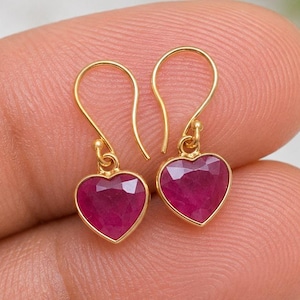 Puede incluir: Un par de pendientes colgantes de tono dorado con piedras preciosas facetadas en forma de corazón de color rosa intenso. Cada pendiente cuelga de un sencillo gancho de alambre dorado curvado, con un engaste de metal pulido que enmarca las piedras.