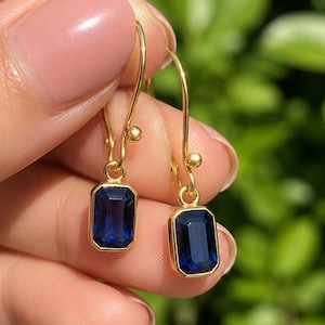 Può includere: Un paio di orecchini pendenti in oro con pietre rettangolari sfaccettate blu intenso incastonate in castoni d'oro lisci. Gli orecchini pendono da sottili ganci in filo d'oro, ciascuno decorato con una piccola perlina d'oro in cima.