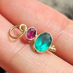 Pode incluir: Um pingente de ouro com uma pequena pedra preciosa rosa de corte oval e uma pedra preciosa verde maior de corte oval, engastadas em uma moldura de ouro. O pingente inclui um anel de junção circular de ouro no topo para fixação em corrente.