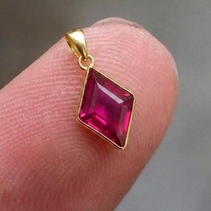 Pode incluir: Um pingente de ouro com uma pedra preciosa vermelha escura em forma de diamante. O pingente é pequeno e delicado, com uma simples argola de ouro para uma corrente. A pedra tem um corte facetado.