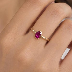 May include: A gold ring featuring a single oval-shaped, deep pinkish-red gemstone set in a smooth bezel setting. The band is thin and polished, showcasing a simple design that highlights the vibrant color and clarity of the central stone.