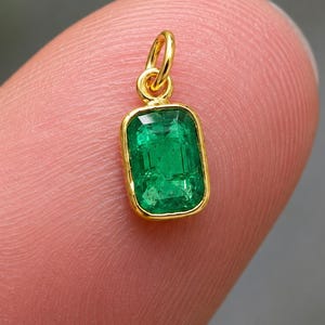 Op de afbeelding: Een kleine rechthoekige smaragdgroene hanger gezet in een geelgouden rand. De levendige groene steen heeft een gefacetteerde snit en is bevestigd aan een eenvoudige gouden springring, wat een delicaat sieraad vormt voor een ketting of armband.