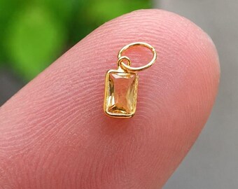 May include: A small, rectangular gold pendant with a light orange gemstone. The pendant has a simple design with a small loop at the top for a chain. The gemstone is a light orange color and is set in a gold frame.