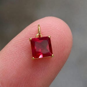 Pode incluir: Um pequeno pendente de pedra preciosa vermelha de corte quadrado, engastado numa armação de metal dourado com quatro garras nos cantos e um pequeno anel circular no topo, apresentando um tom carmesim vibrante e superfície facetada.