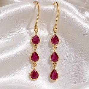 May include: A pair of gold drop earrings featuring three vertically aligned, teardrop-shaped, deep pink gemstones suspended from a curved gold hook. The earrings have a polished metallic finish and a delicate, articulated design that allows for movement.