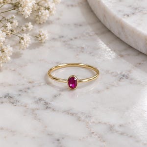 May include: A thin gold ring featuring a single, small, oval-shaped, vibrant pinkish-red gemstone set in a smooth bezel setting. The band is polished and slender, creating a delicate appearance for this piece of fine jewelry.